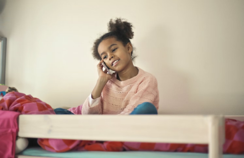 A little girl sits on a bed and holds a cell phone up to her ear.